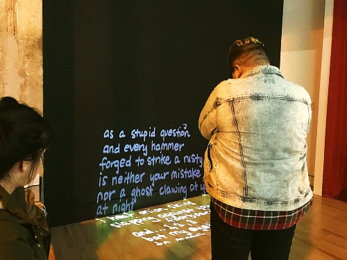 A gender non-conforming person with short, bleached hair and glasses reads Jaye Sablan's poem called Witness. The poem is being project onto a wall at the Hedreen Gallery. Jaye contributed this poem for artist E.T. Russian's Double Clear exhibit (01/09/19, Seattle WA).