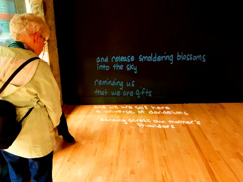 An older White woman, short hair, and glasses reads Jaye Sablan's poem called Tronkun At'but. The poem is being project onto a wall at the Hedreen Gallery. Jaye contributed this poem for artist E.T. Russian's Double Clear exhibit (01/09/19, Seattle WA).